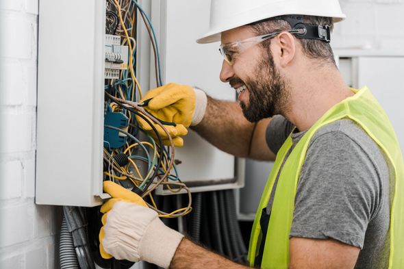 Electrician in hard hat and safety vest, working on electrical panel, smiling.