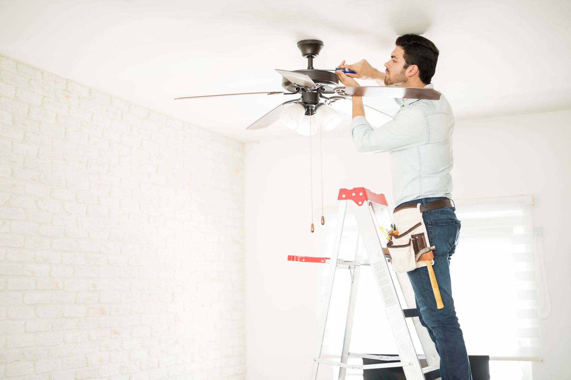 Man on a ladder installing a ceiling fan in a white room.