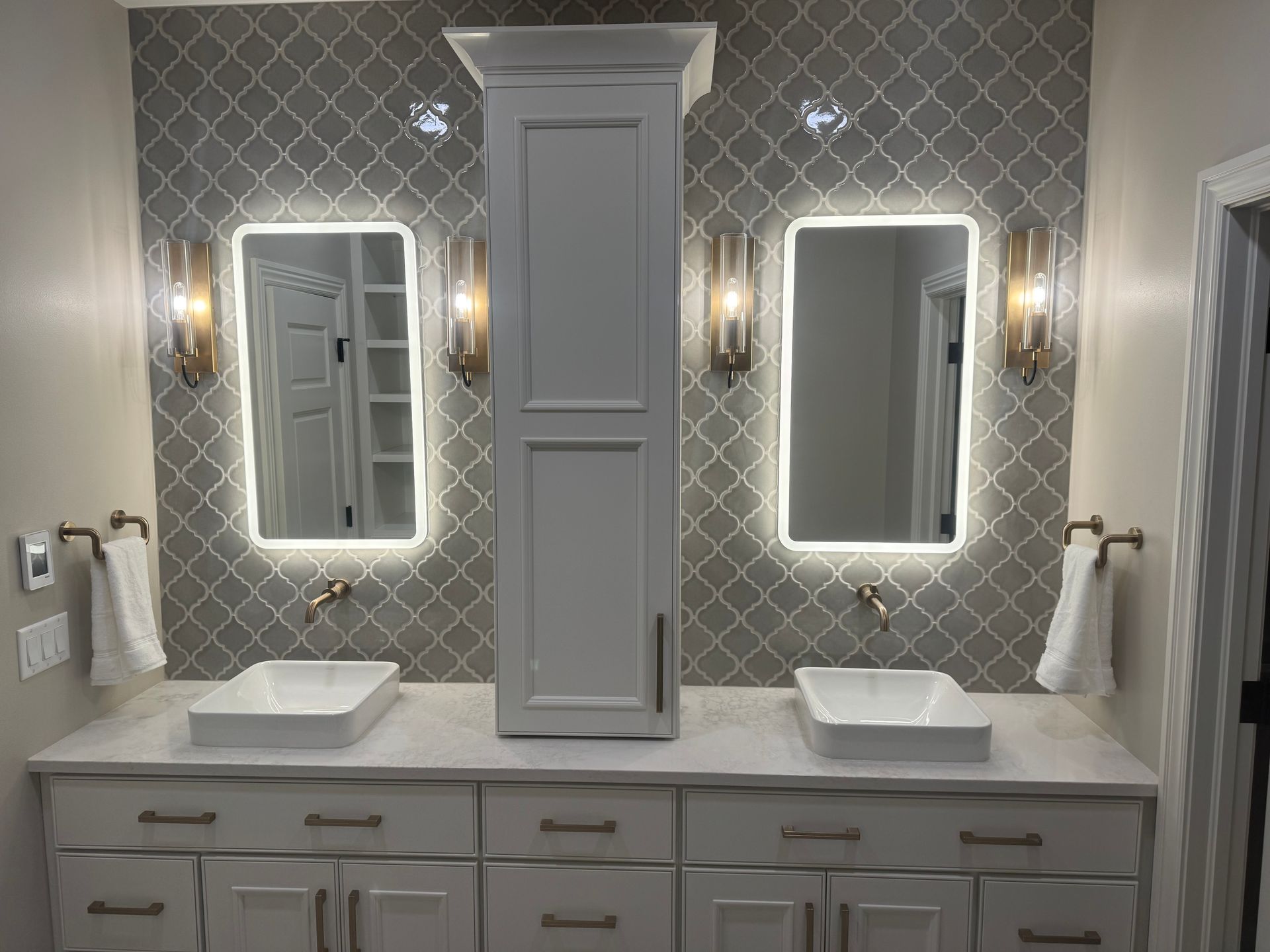Bathroom with two lighted mirrors, white cabinets, grey tile backsplash, and vessel sinks.
