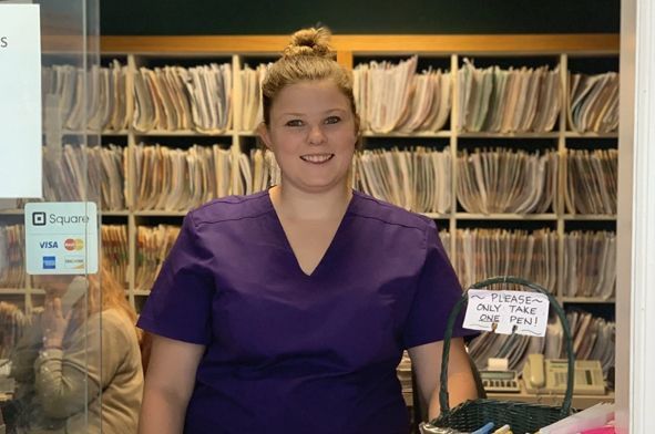 A woman in a purple scrub top is standing in front of a row of shelves.