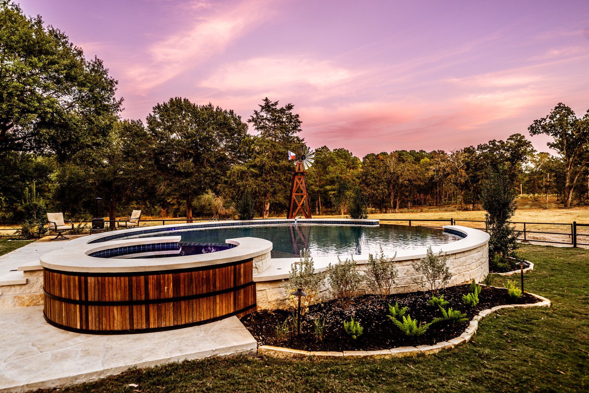 Pool and spa with wooden accents, surrounded by greenery and set against a sunset sky.