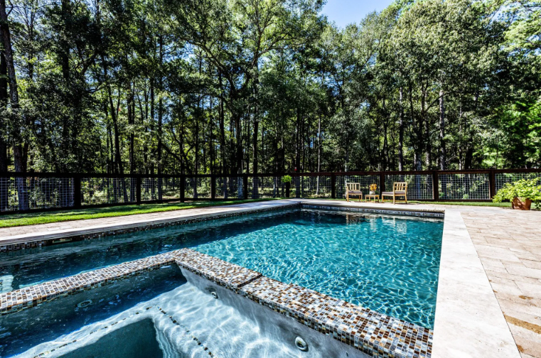 Pool with clear turquoise water and stone steps. Green lawn, dark fence, and tall trees.