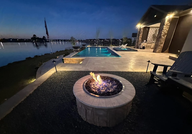 A fire pit on a patio next to a pool and lake at dusk.