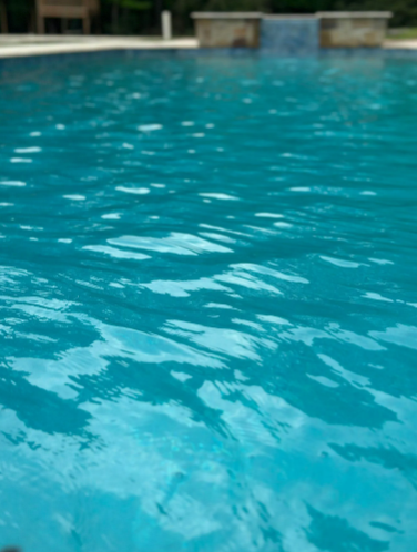 Close-up of a blue swimming pool water surface with ripples, blurred background of pool edge and building.