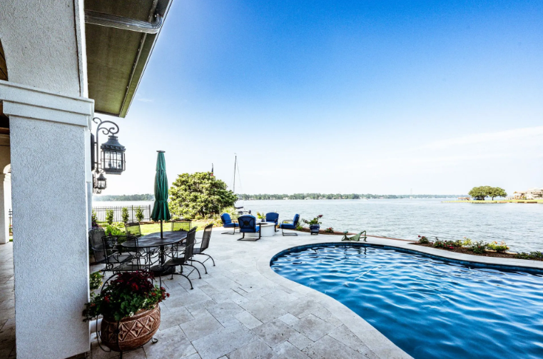 Poolside patio overlooking a lake, with a pool, chairs, and umbrella under a bright blue sky.