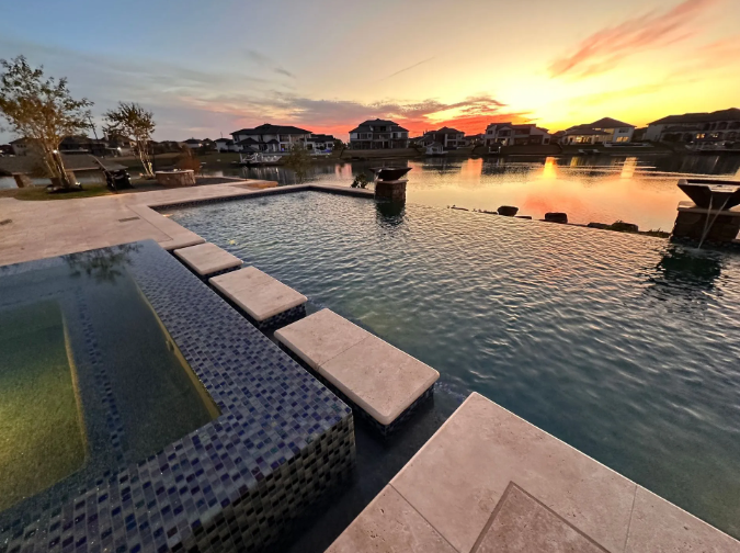 Sunset over a pool and lake with homes in the background. The pool has a dark blue tiled area and tan borders.