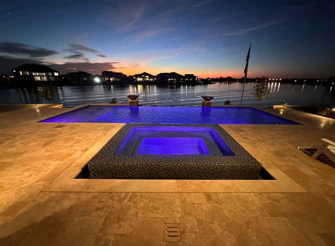 Night view of a pool and spa with blue lighting. Sunset over water in the background, with houses lit up along the shoreline.