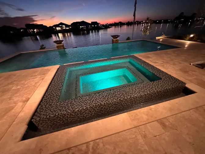Pool with attached hot tub, set against a lake at dusk. Blue and tan tones dominate.