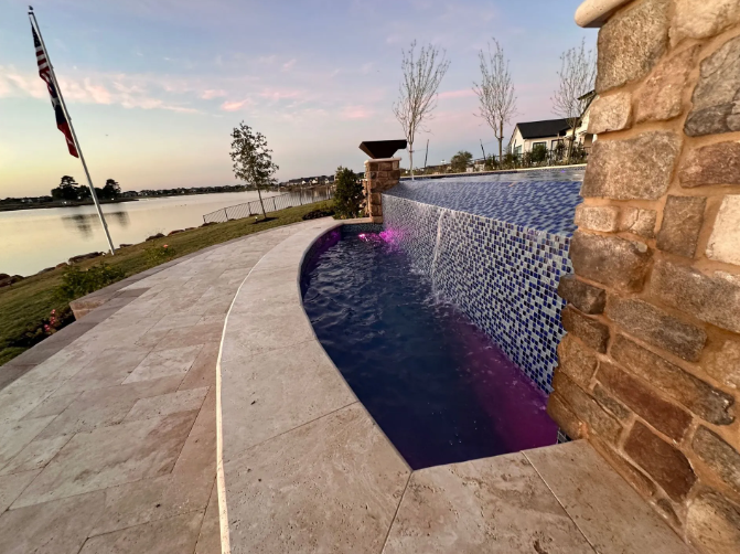 Pool with a stone wall and waterfall overlooking a lake at sunset.