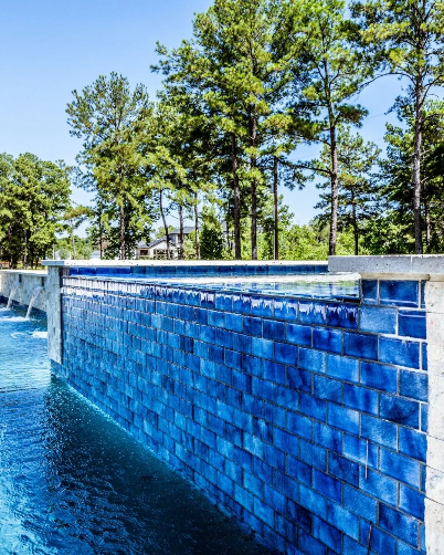Blue tiled wall with water cascading into a pool. Tall trees and a clear blue sky in the background.