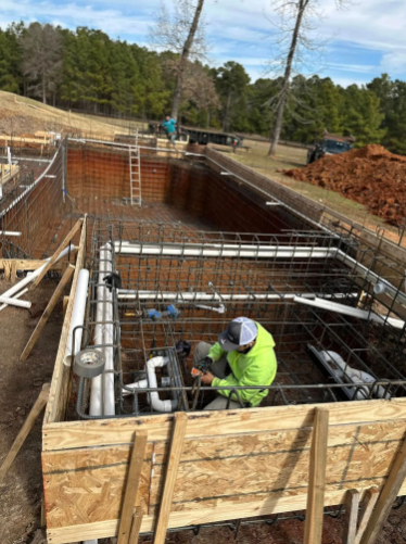 Construction worker installing pipes inside a pool form. Rebar and wooden framing surround the pool's excavated space.