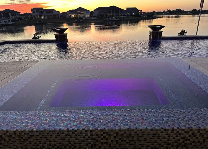 A square pool with purple lights at sunset, overlooking a lake with houses.