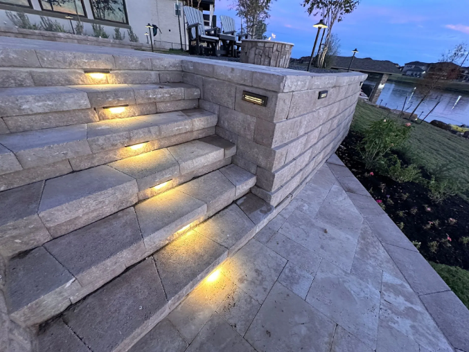 Stone steps with integrated lighting lead down to a patio beside a lake.