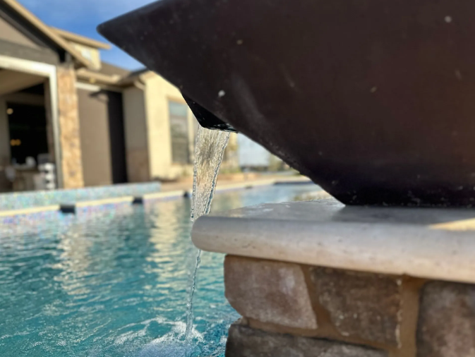 Water pours from a dark vessel into a swimming pool with a stone and brick border; a house is in the background.