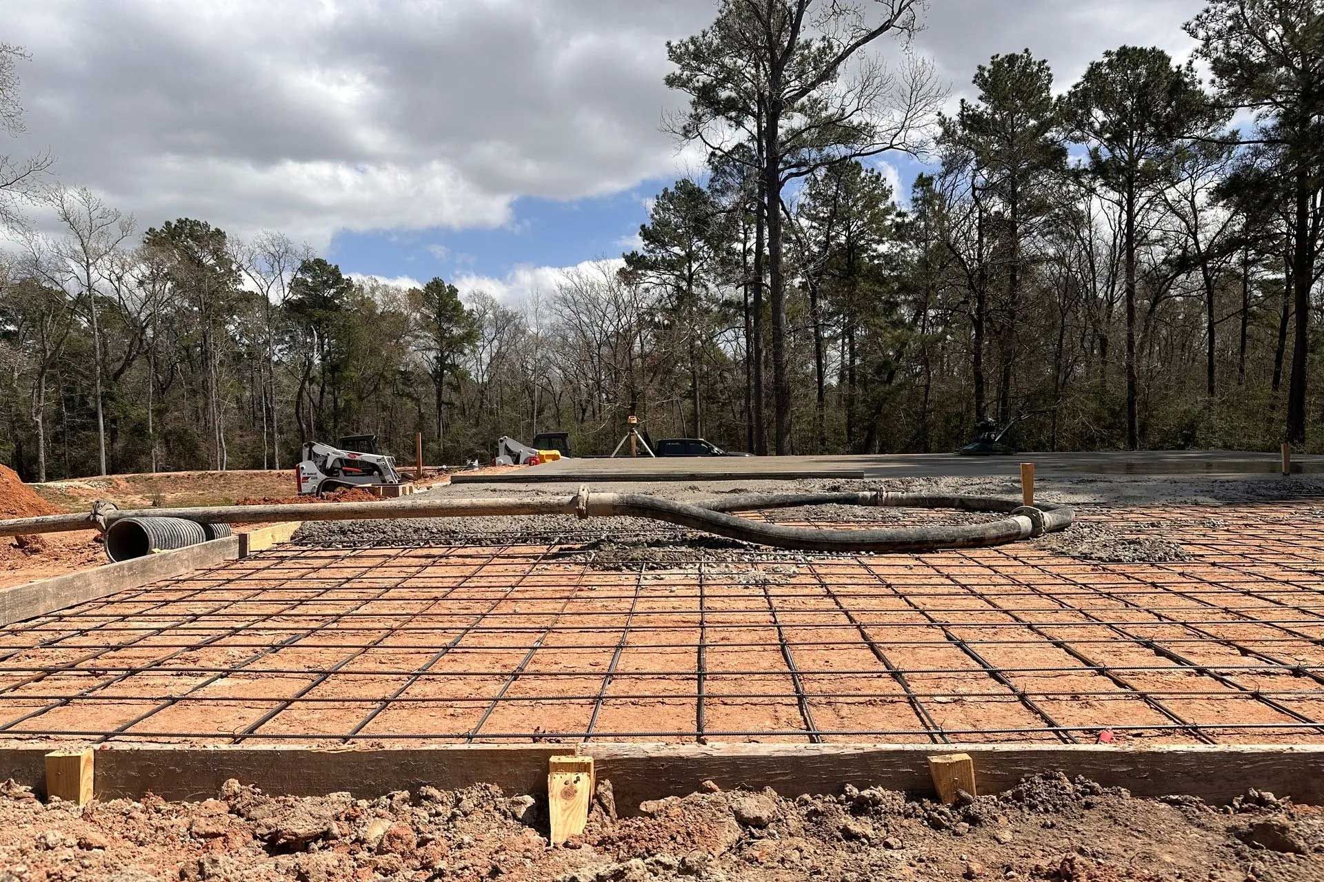 Construction site with a concrete foundation frame, rebar grid, and surrounding dirt. Forest background.