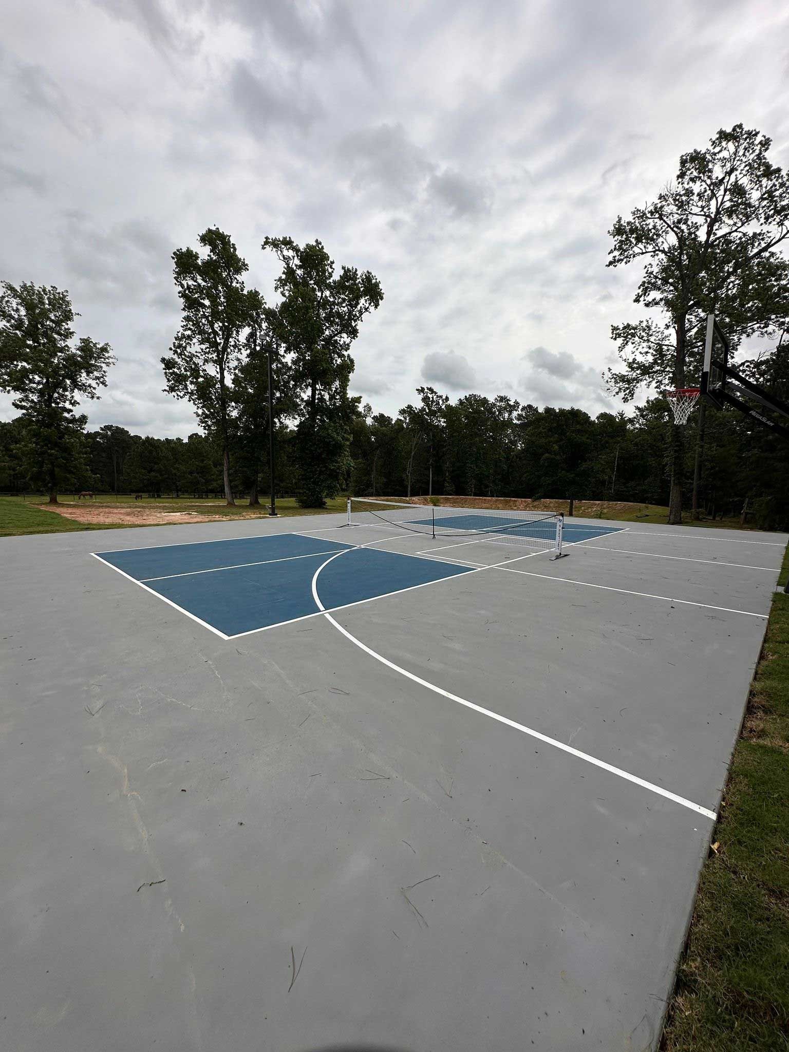 Outdoor basketball court with blue key, gray pavement, and trees under cloudy sky.