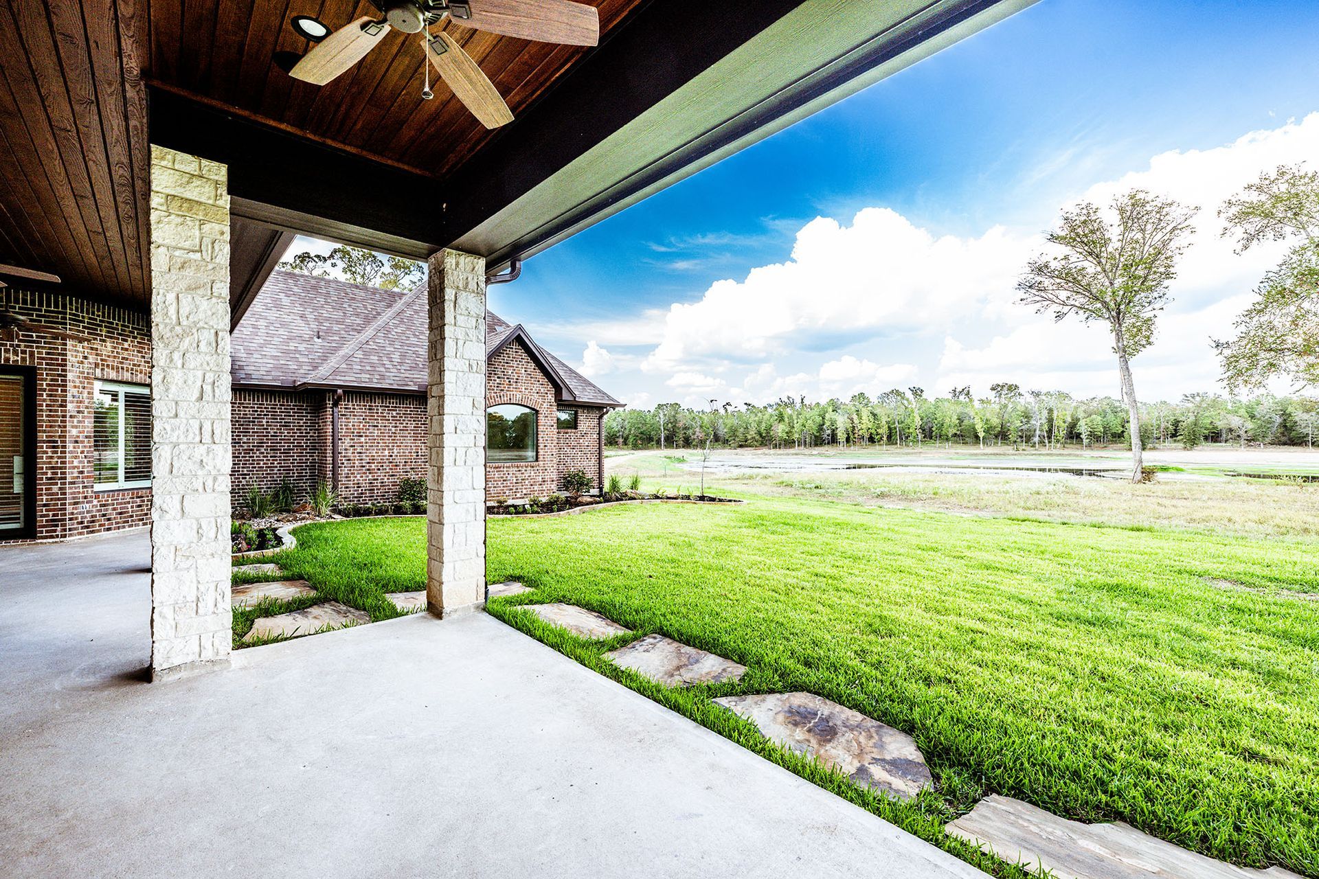 Patio overlooking a green lawn and small cabin. Stone path, blue sky, and a ceiling fan.