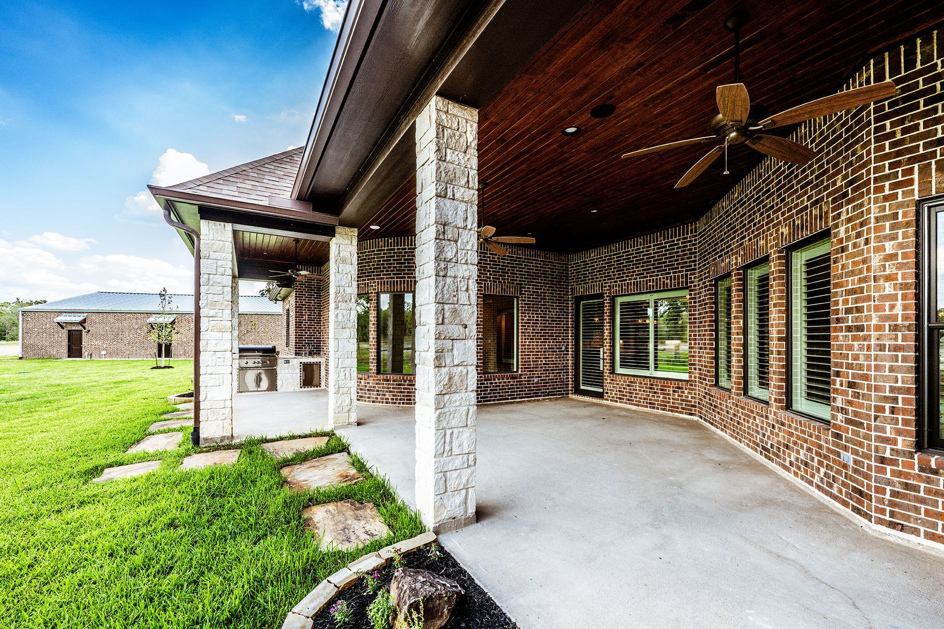 Covered patio with stone pillars and brick walls, green grass, and blue sky.