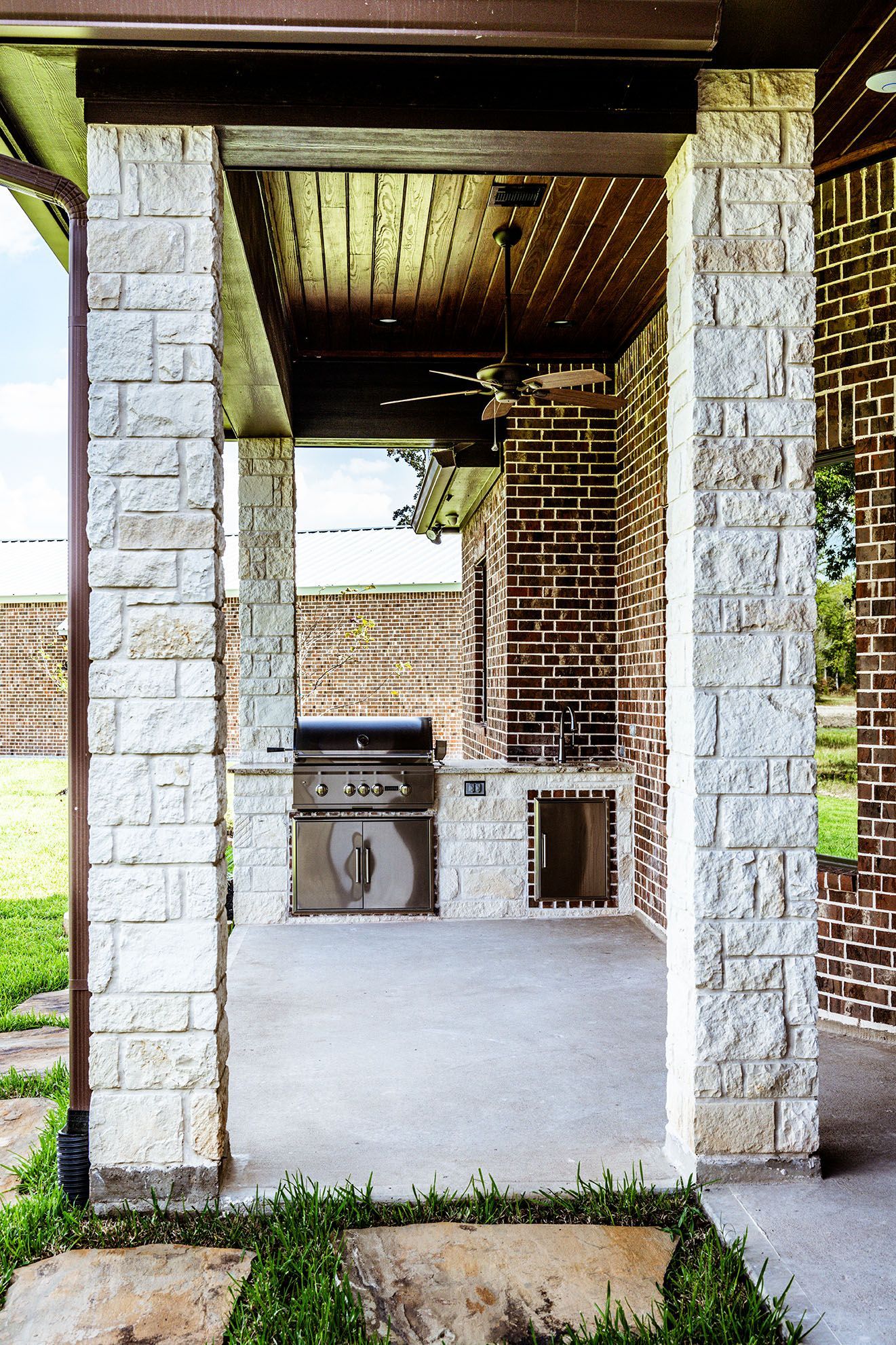 Outdoor kitchen with stone pillars, a grill, sink, and brick backdrop.