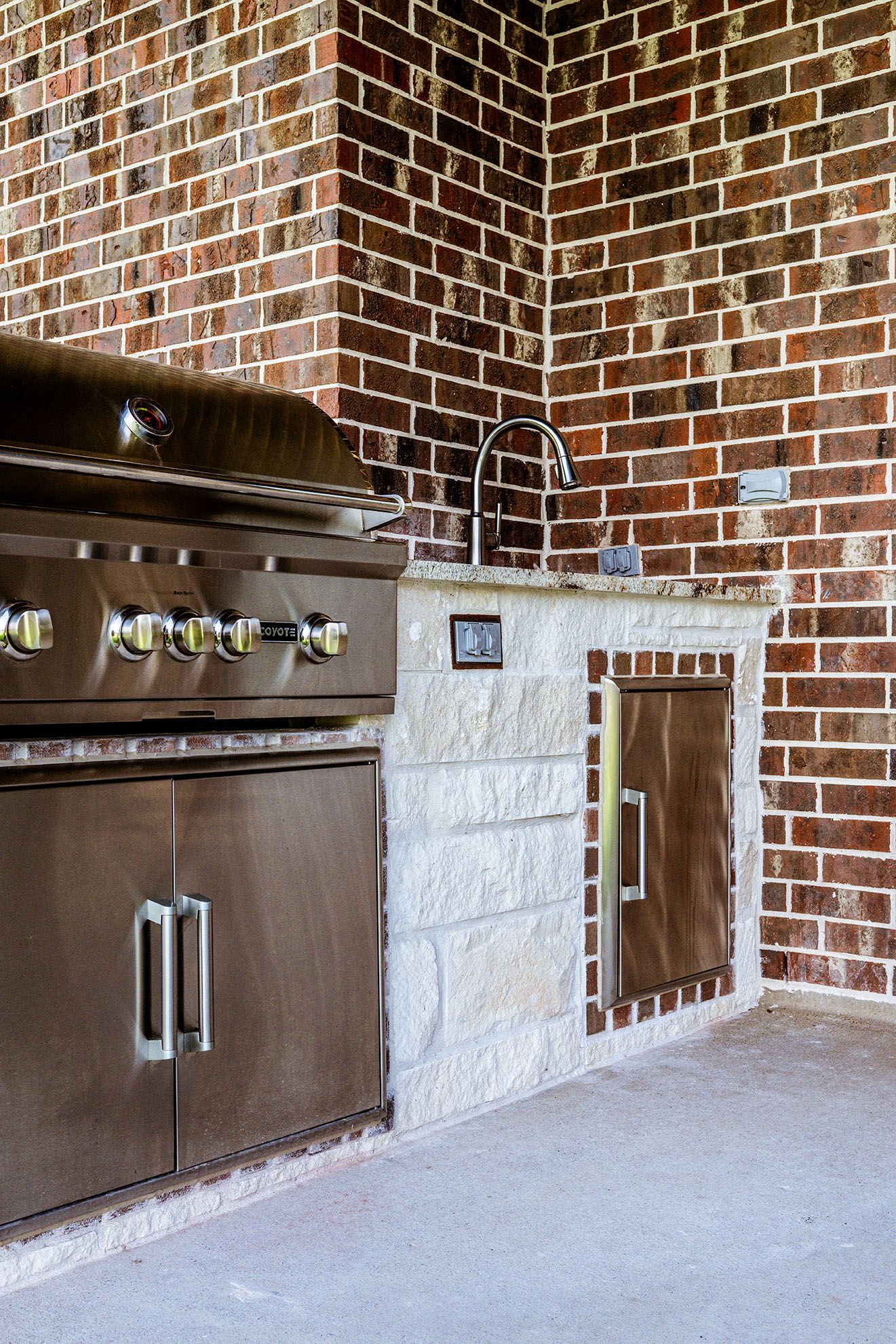 Outdoor kitchen with stainless steel grill, brick walls, and a stone countertop with a sink.