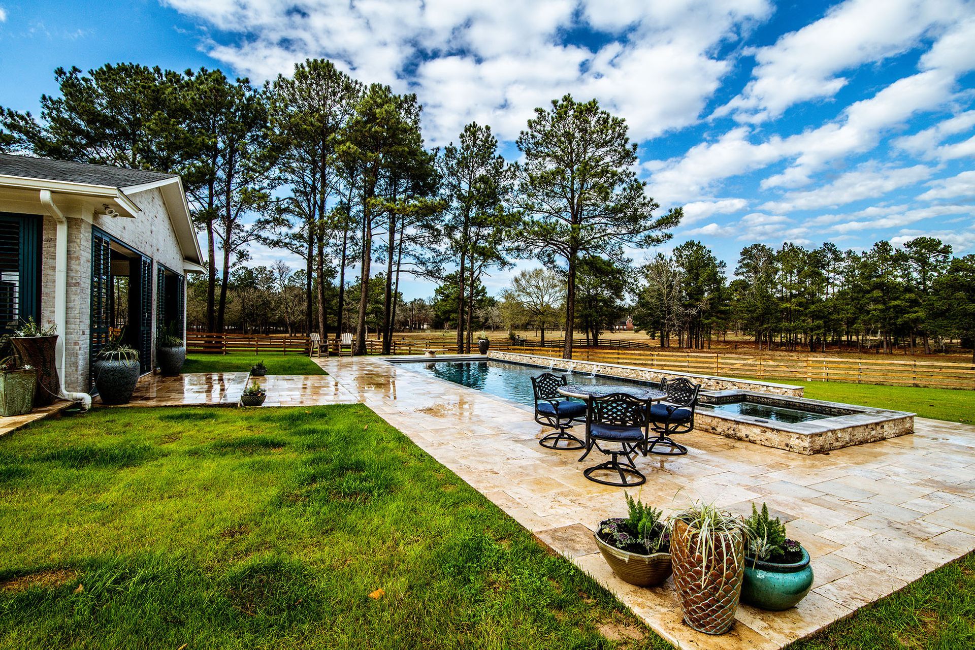 Backyard with pool, patio, house, and trees under a blue sky with clouds.