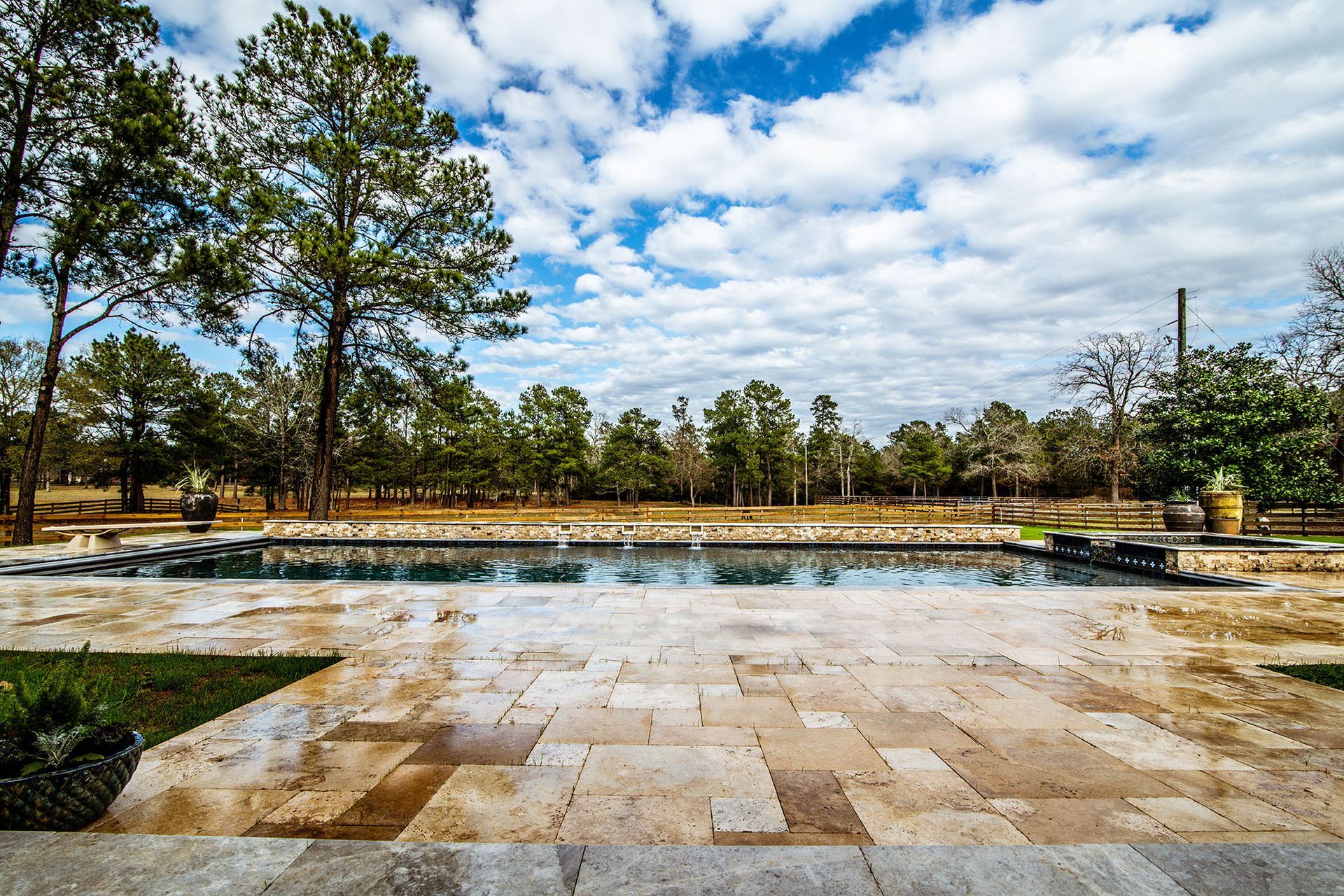 A rectangular pool with clear water, surrounded by stone patio. Trees and a cloudy blue sky in the background.