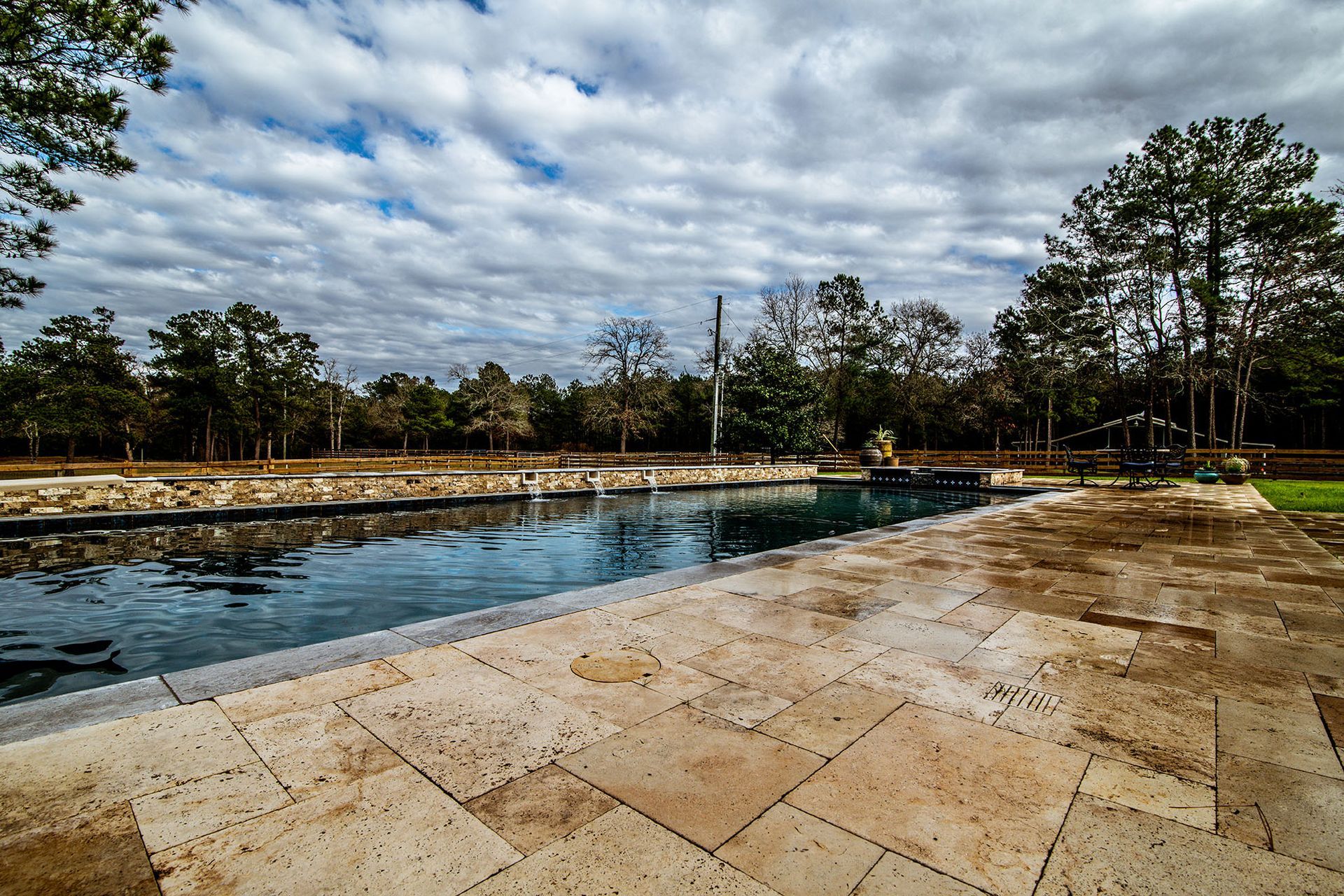 Swimming pool surrounded by stone patio, trees, and cloudy sky.