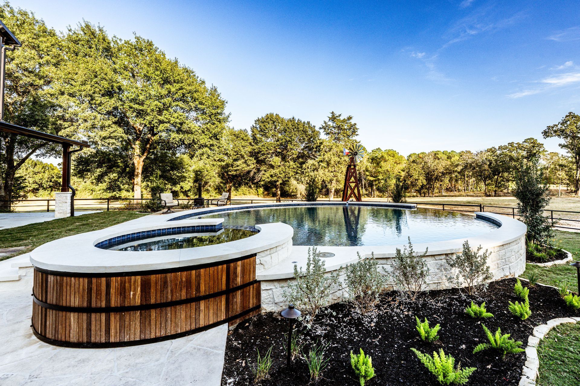 Pool with a wooden hot tub, concrete and stone surround, and lush greenery under a blue sky.