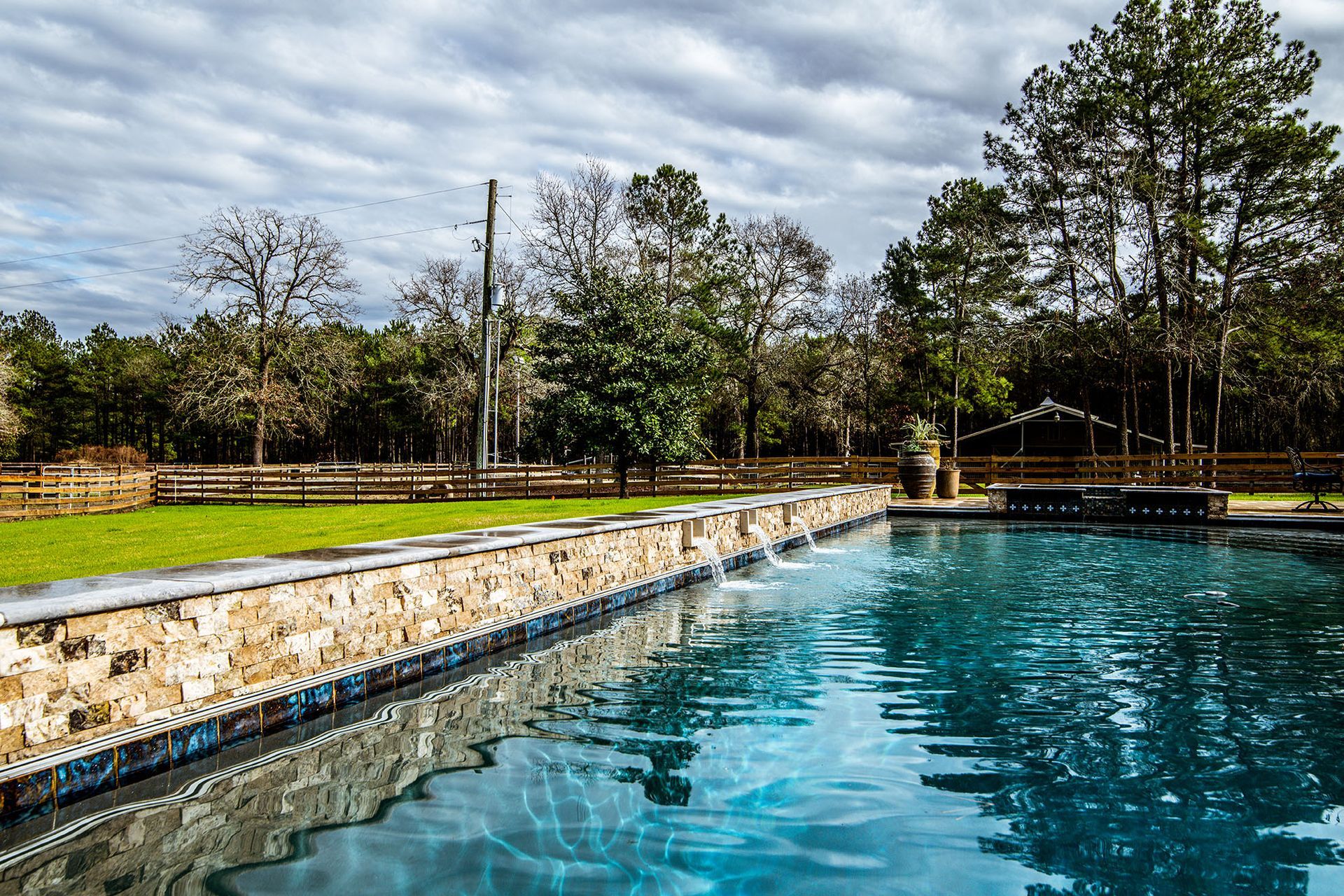 Stone lined pool with blue water, green lawn, wooden fence, trees, and cloudy sky.