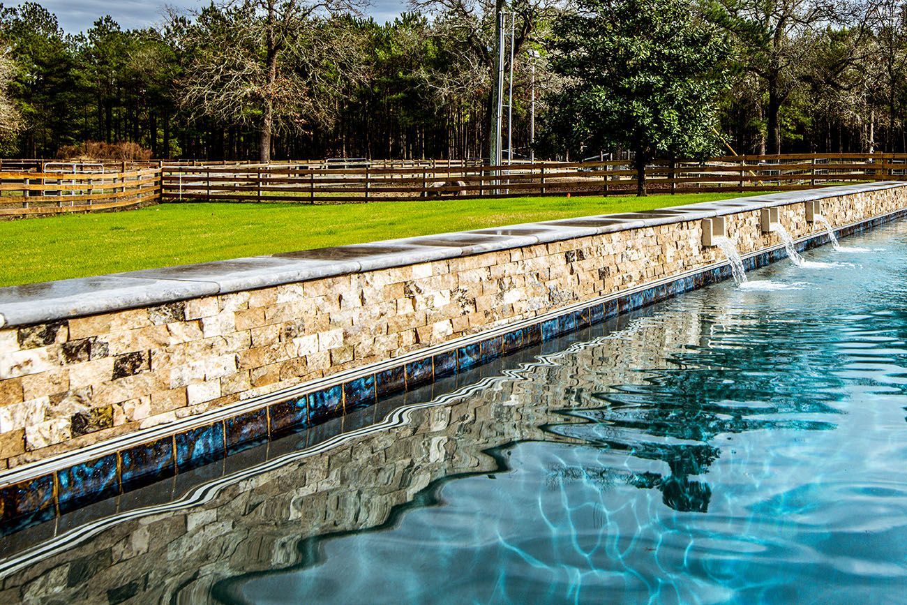 Pool with stone edging and water jets, next to a grassy lawn and wooden fence, with trees in the background.