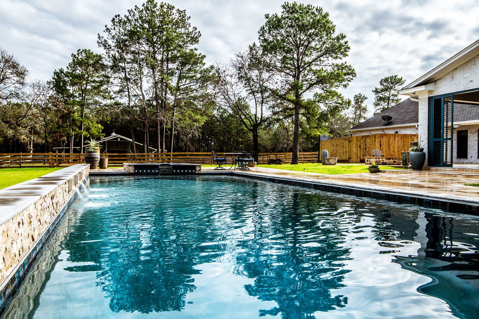 Swimming pool with clear blue water, surrounded by grass and trees, and a house in the background.