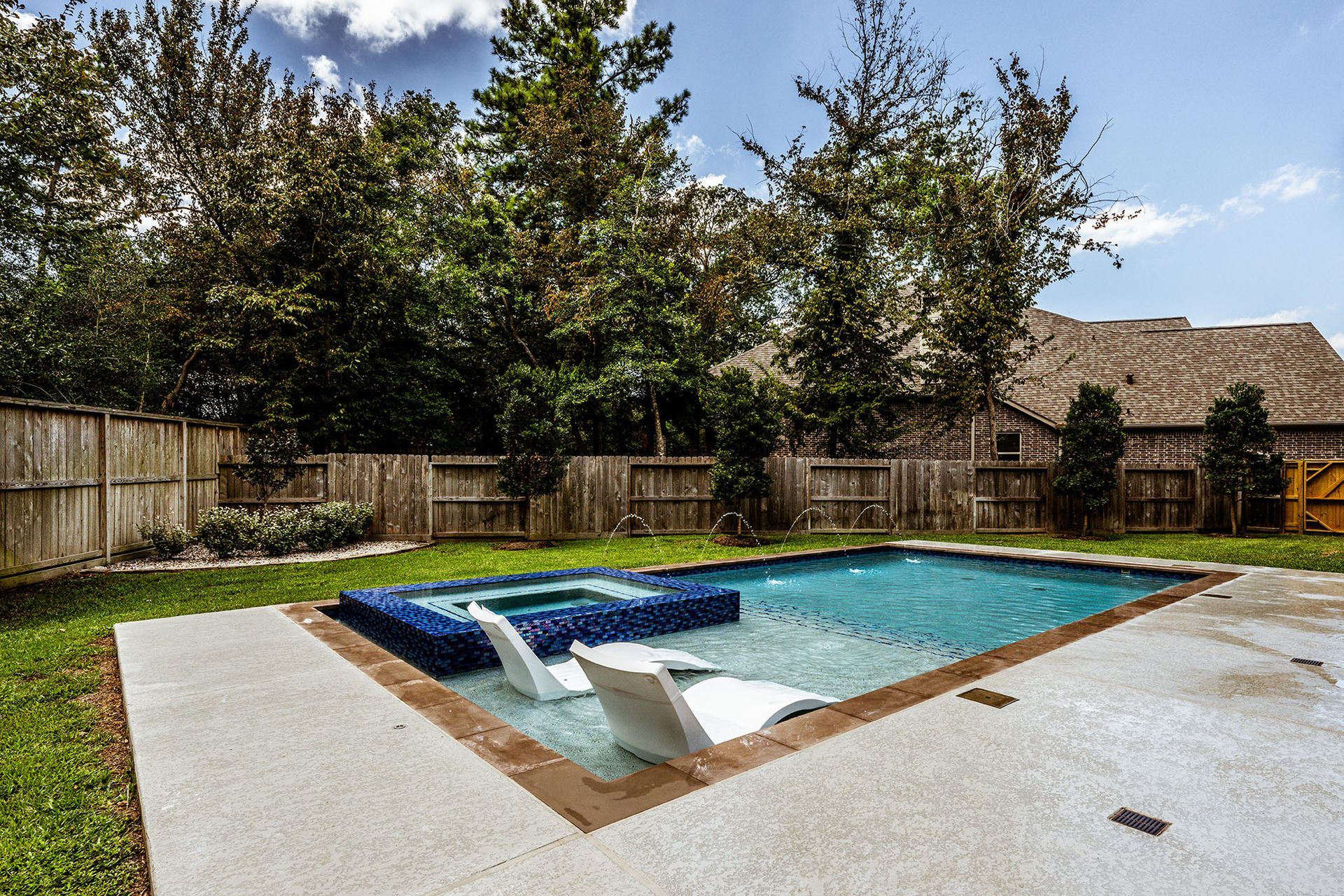 Backyard pool with spa, lounge chairs, concrete patio, and wooden fence. Trees and blue sky in background.