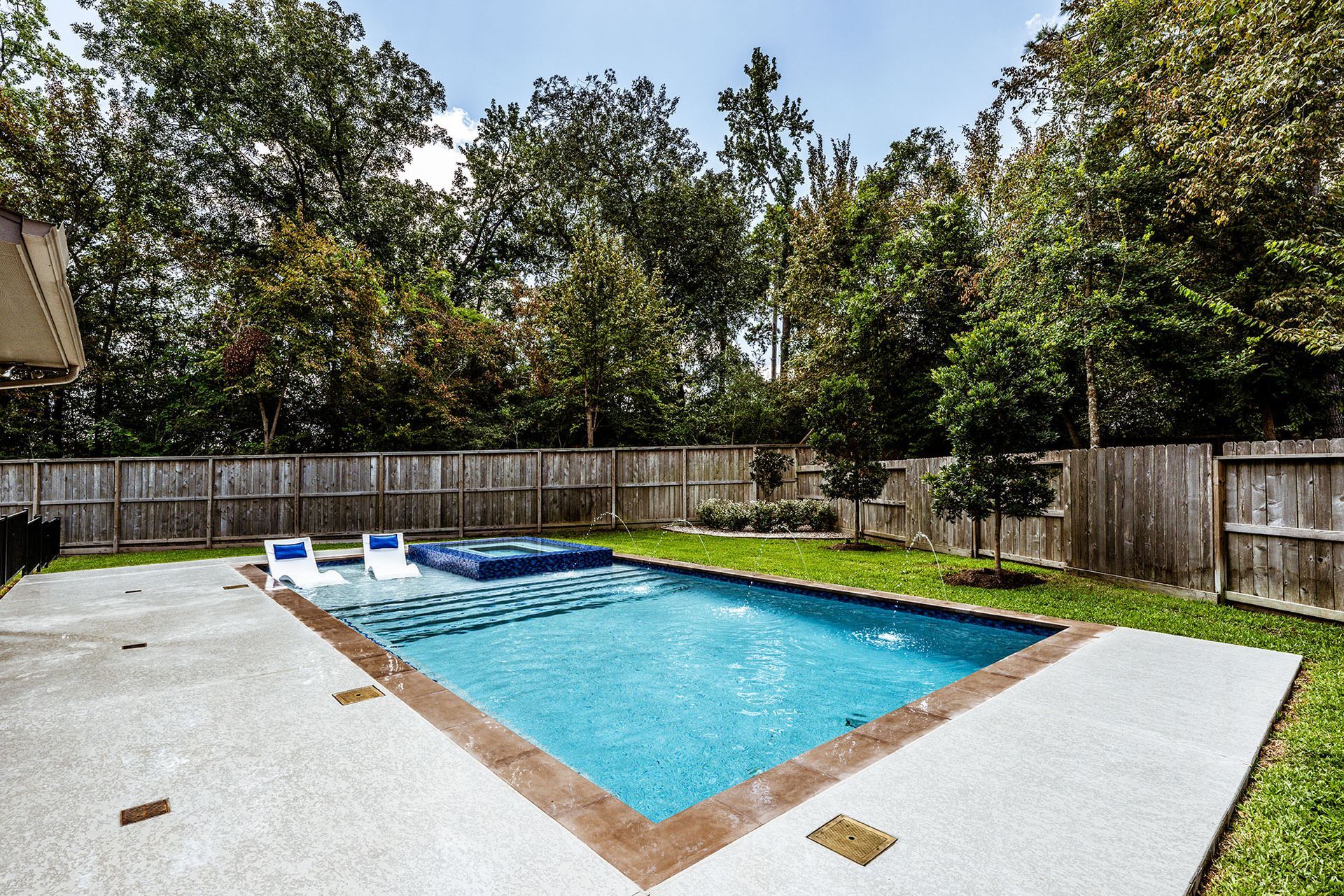 Backyard pool with spa, surrounded by concrete patio and wooden fence trees in background.