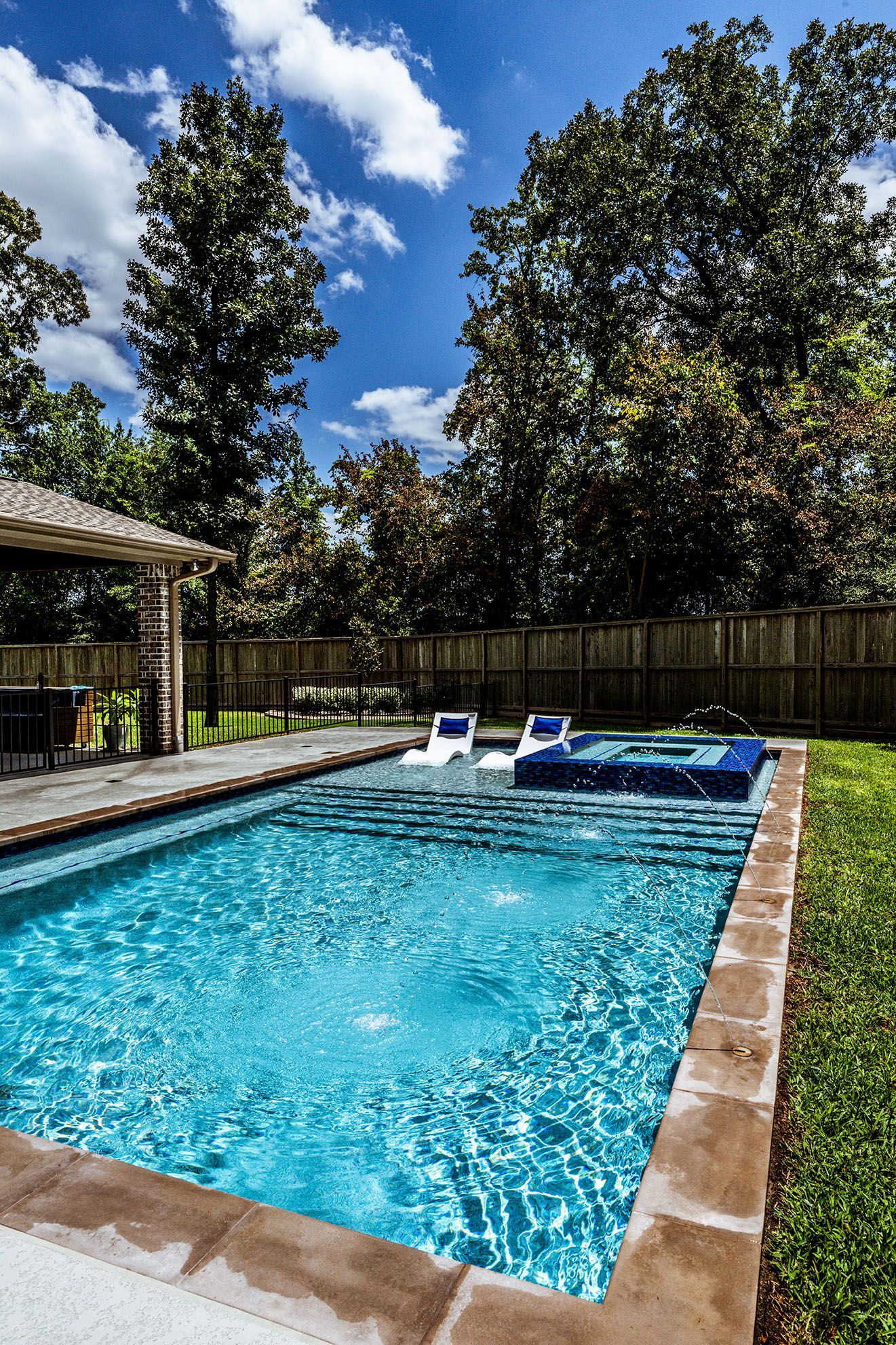 Rectangular swimming pool with bubbling water, blue water, and two white lounge chairs. Trees and a sunny sky.