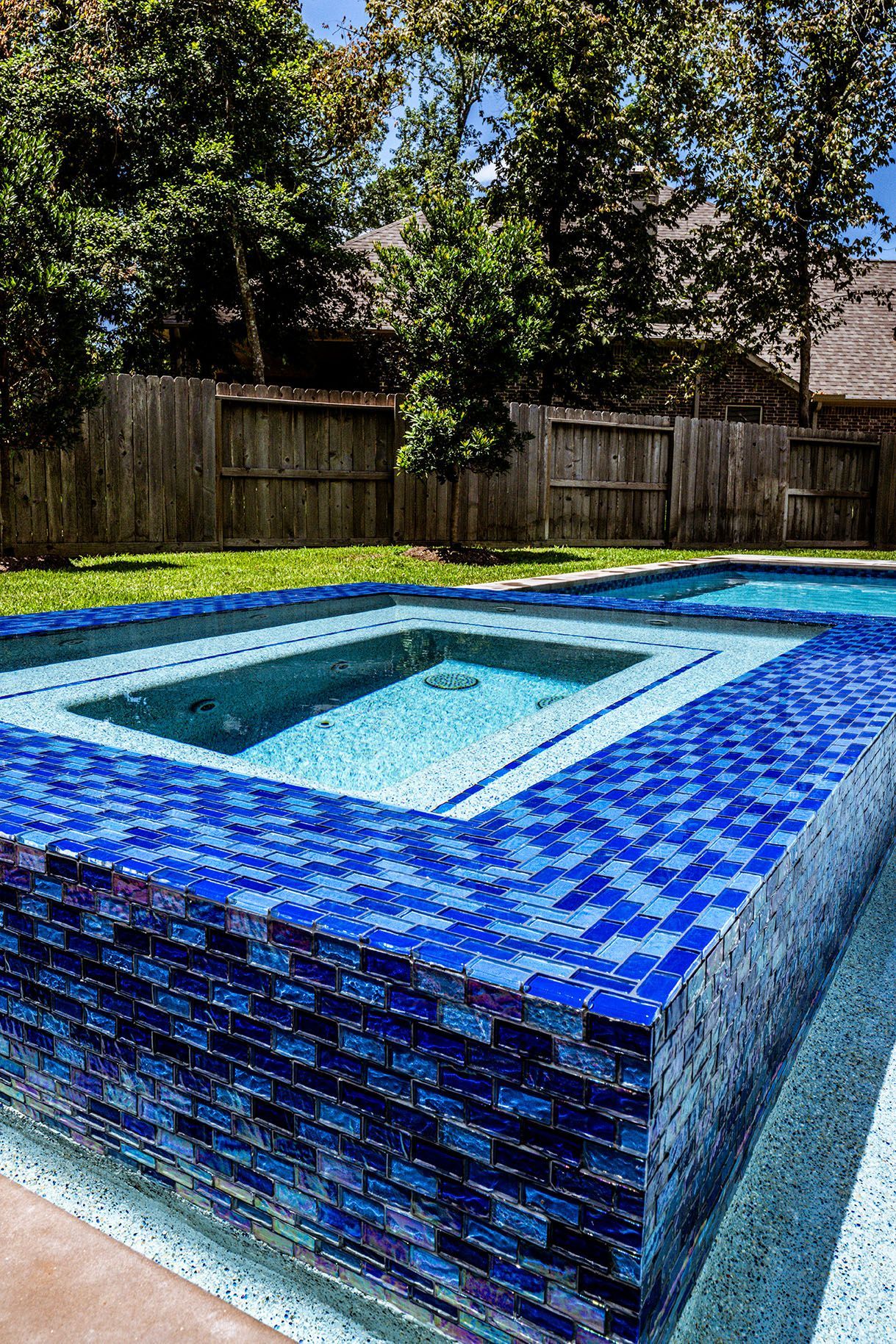 Blue-tiled pool and spa in backyard, surrounded by a wooden fence and trees.