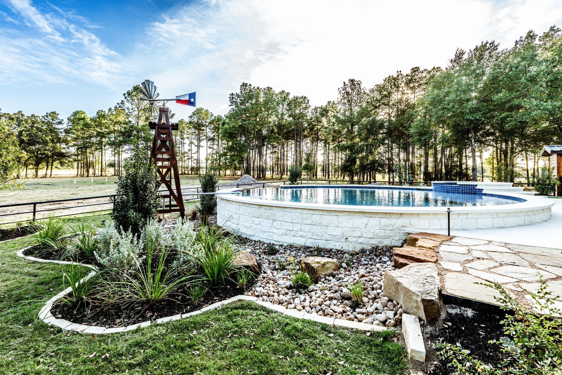 Pool with white stone border, windmill with Texas flag, surrounded by trees and landscaping.