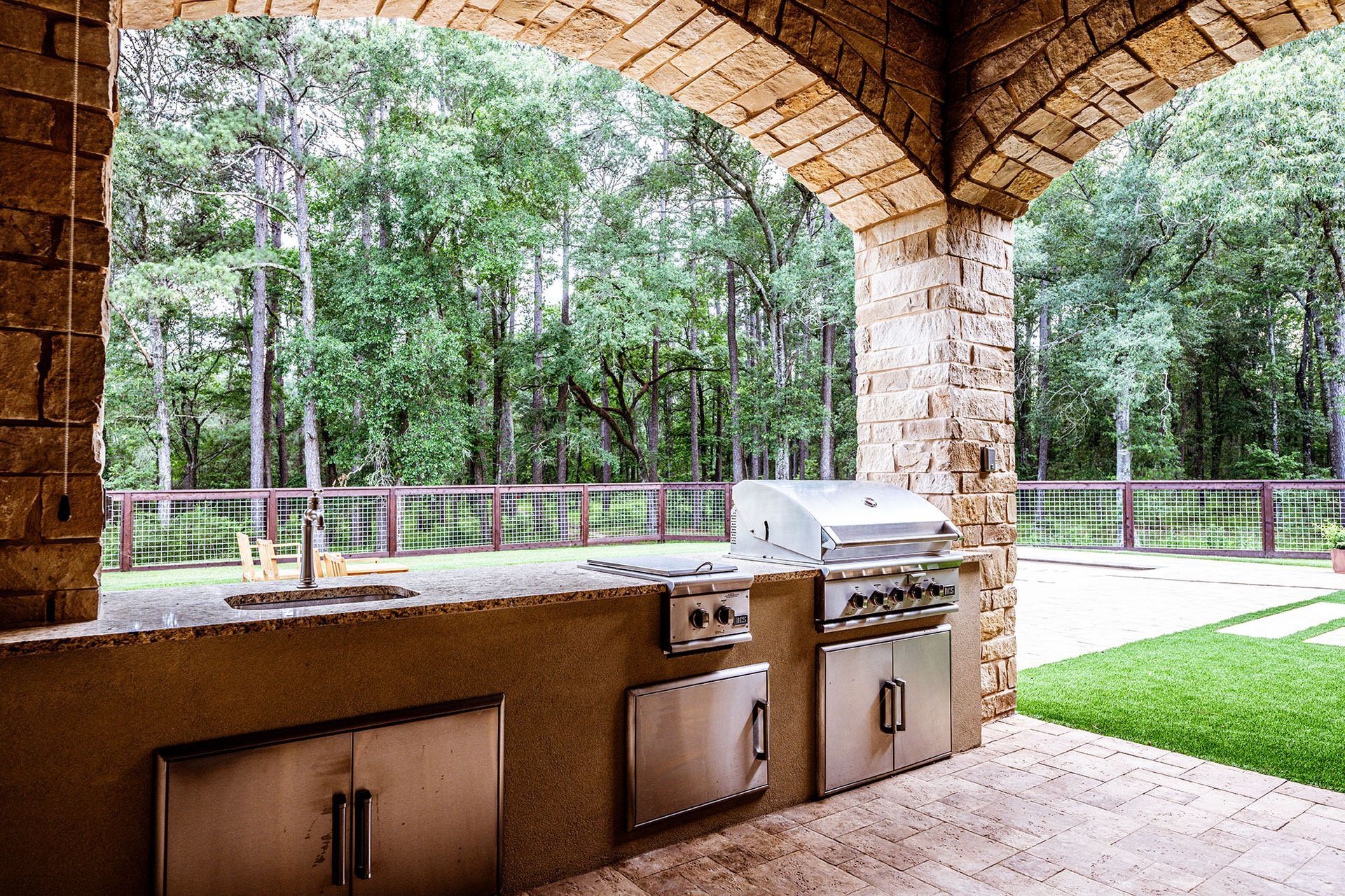 Outdoor kitchen under a stone archway with a grill, sink, and stainless steel cabinets overlooking a wooded area.
