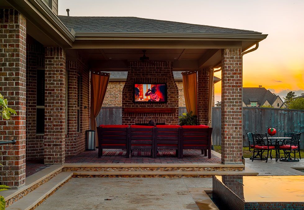 Outdoor brick patio with TV, seating, and curtains sunset in background.