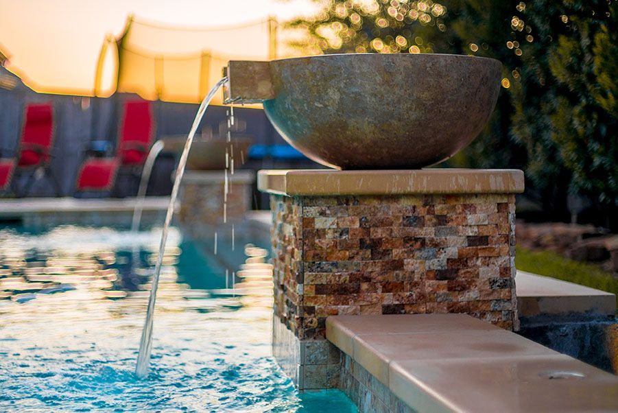 Water fountain with a bowl shaped feature, cascading into a pool. Brick and stone detail, sunny outdoor setting.