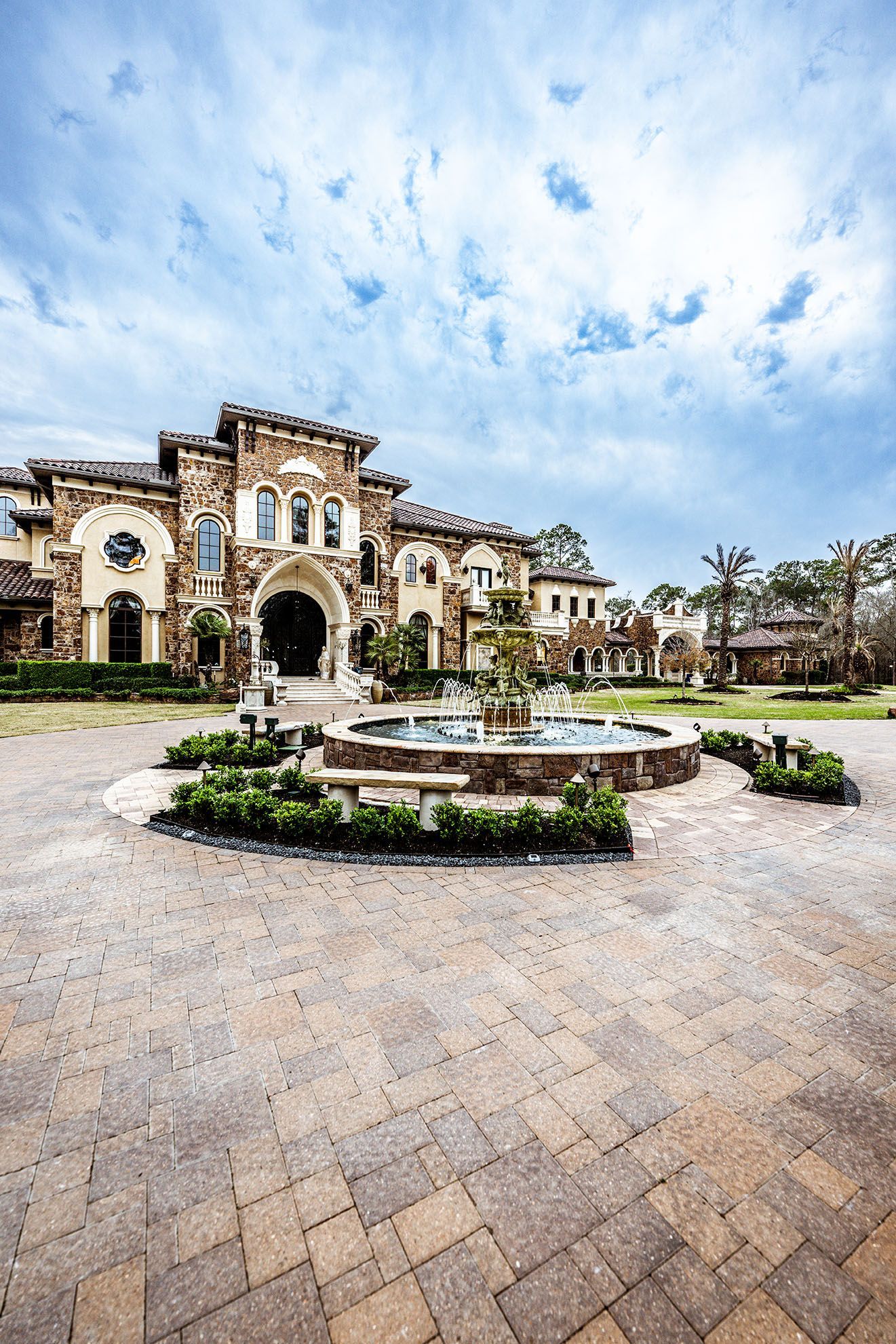 Luxurious stone mansion with fountain in front; circular brick driveway under cloudy sky.