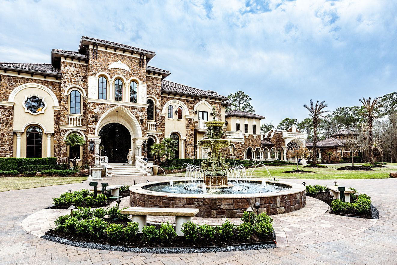 Large stone mansion with fountain and manicured landscaping under a cloudy sky.
