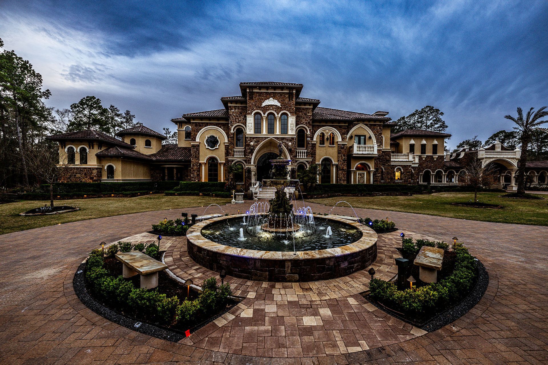 Mansion with a fountain in front. Tan stone exterior, arched windows, and dark sky.