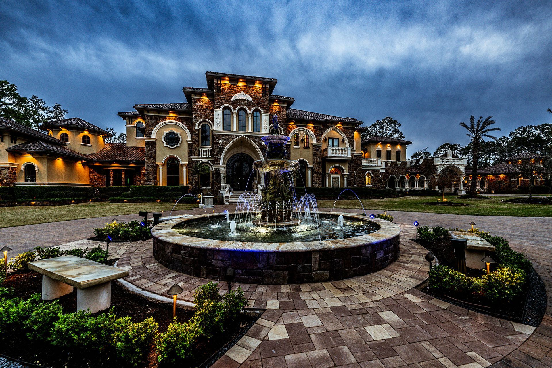 Grand mansion with fountain in front evening view with cloudy sky and lights.