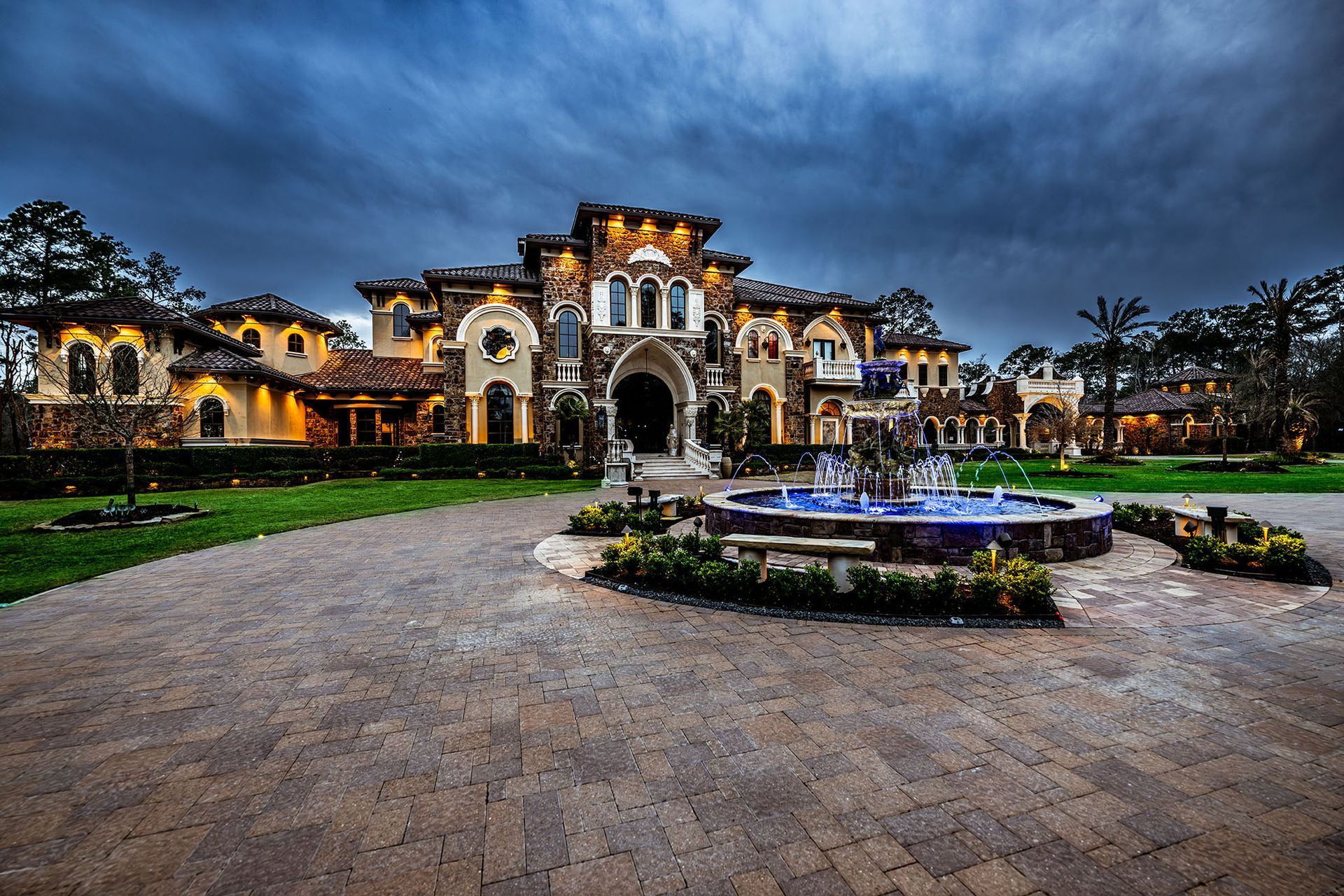 Ornate stone mansion with a fountain on a brick driveway, under a cloudy evening sky.
