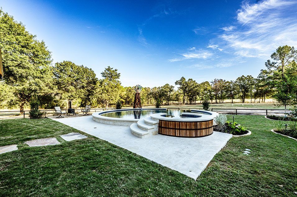 Backyard with pool and hot tub surrounded by green grass and trees. Blue sky overhead.