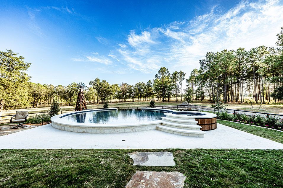 Outdoor swimming pool and hot tub surrounded by trees under a blue sky.