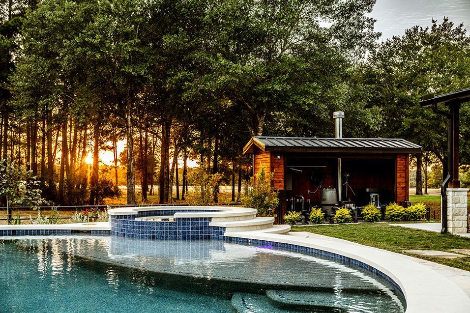 Poolside view with pool, hot tub, small wooden building with chimney, and trees at sunset.