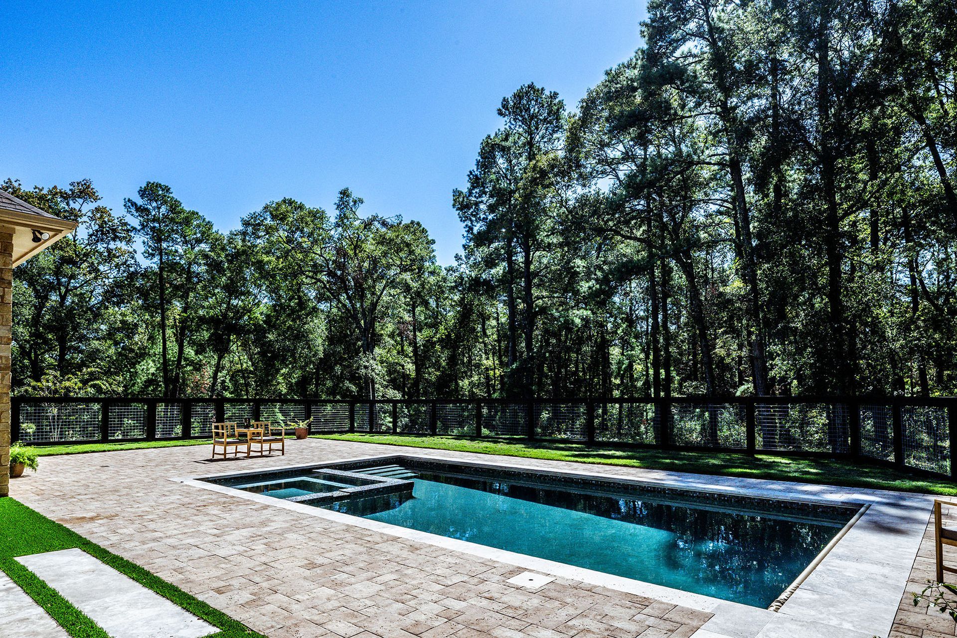 Rectangular pool with clear water, surrounded by stone patio and fenced backyard with trees.
