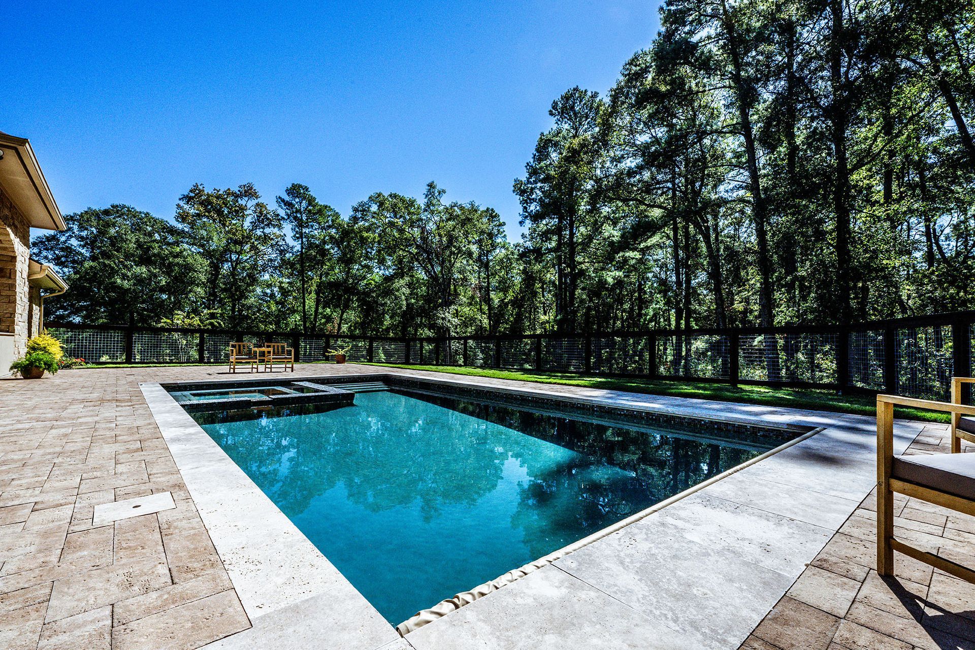Rectangular pool with turquoise water, stone patio, and view of green trees under blue sky.