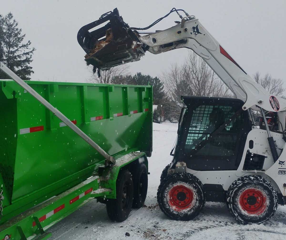 Bobcat loader emptying debris into a green trailer in a snowy outdoor setting.